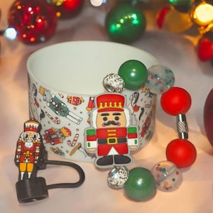 May include: A Christmas-themed charm bracelet with red, green and clear beads. The bracelet features a nutcracker charm and a charm with a white rhinestone setting. The bracelet is displayed on a white cup with a nutcracker pattern.