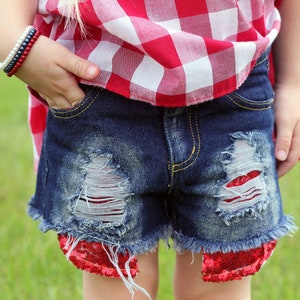 May include: Distressed denim shorts with red sequin trim. The shorts have a frayed hem and are decorated with red and white patches.