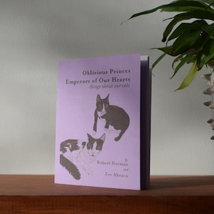 May include: A book titled "Oblivious Princes Emperors of Our Hearts things about our cats" is propped up on a wooden surface. The book has a lavender cover with illustrations of cats. The book is by Robert Norman and Eve Ahearn.