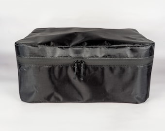 Puede incluir: Un cubo de almacenamiento negro con cremallera hecho de tela resistente al agua. El cubo tiene una cremallera con dos tiradores.
