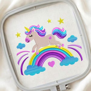 Puede incluir: Diseño bordado de un unicornio saltando sobre un arcoíris. El unicornio tiene un cuerpo rosa, un cuerno amarillo y una melena y cola coloridas. El arcoíris es en tonos de amarillo, azul, morado y rosa, con nubes y estrellas.