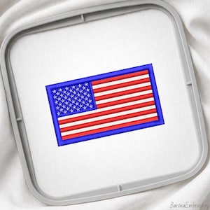 May include: An embroidered patch of the United States flag featuring a blue canton with white stars and alternating red and white horizontal stripes, secured within a grey plastic embroidery hoop on a white fabric background.