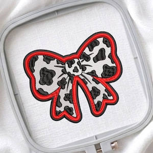 May include: An embroidered bow patch featuring a white and black cow print pattern. The bow is outlined with a thick, vibrant red border and is secured within a light gray plastic embroidery hoop against a white fabric background.