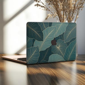 Può includere: Un computer portatile con una cover protettiva caratterizzata da un motivo botanico a foglie in verde ottanio scuro e verde salvia. Il design presenta sottili linee color oro che delineano le venature delle foglie e un logo Apple nero.