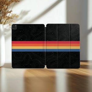 May include: A black tablet case featuring a white topographic map pattern with scattered white dots. A horizontal stripe design runs across the center in red, orange, and blue. The case is shown in an open, unfolded position with a camera cutout.