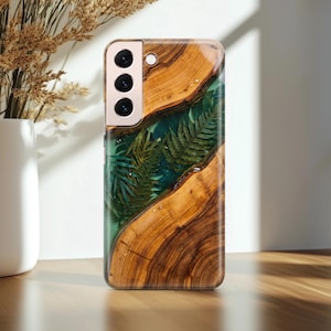 Könnte beinhalten: Eine Handyhülle aus Holz mit einem einzigartigen Design. Die Hülle hat ein natürliches Holzmaserungsmuster mit einem klaren Harzeinleger mit grünen Farnblättern. Die Handyhülle hat ein Kameramodul mit drei Objektiven und einem Blitz.