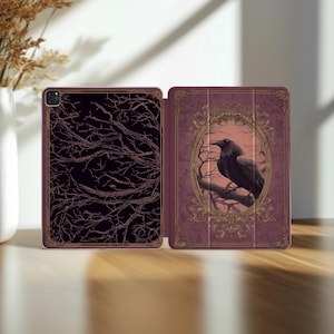 May include: A tablet case with a dark burgundy cover. The left side features a black and bronze tree branch design. The right side has a framed image of a raven perched on a branch, set against a dusky pink background.