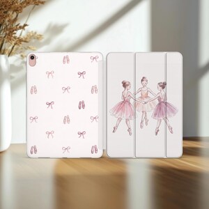 Op de afbeelding: Een witte tablethoes met een herhalend patroon van roze balletschoenen en strikjes op de achterkant, en een illustratie van drie ballerina's in roze tutu's die elkaars hand vasthouden op de drievoudige voorkant met camera-uitsparing.