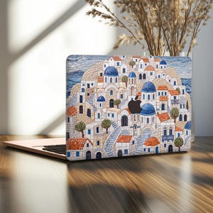 May include: A laptop skin featuring a mosaic-style illustration of a Mediterranean hillside village with white buildings, blue domed roofs, and terracotta-colored accents. The design includes stone staircases, small trees, and a black Apple logo.