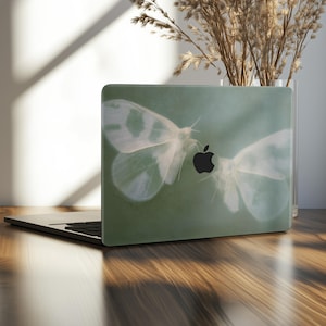 May include: A laptop with a green cover featuring a design of two white moths and a black apple logo. The moths have translucent wings with white spots. The laptop is on a wooden surface.