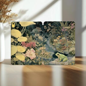 Könnte beinhalten: Eine iPad-Hülle mit einer skurrilen Gartenszene mit einem grünen Frosch, Pilzen und verschiedenen Blumen. Die Hülle ist geöffnet und zeigt das Innendesign. Die Farbpalette umfasst Grün-, Rosa- und Gelbtöne.