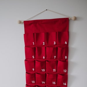 Puede incluir: Un calendario de adviento de pared hecho de tela roja con veinticuatro bolsillos numerados y un saco rojo a juego con cord&oacute;n y un gr&aacute;fico de lazo blanco. El calendario cuelga de una varilla de madera con una cuerda y el saco est&aacute; sobre una silla rosa.