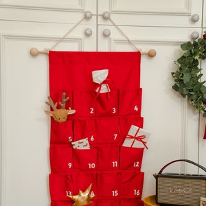 Puede incluir: Un calendario de adviento de tela roja colgado de una varilla de madera, con veinticuatro bolsillos individuales numerados en blanco. Los bolsillos contienen varios objetos peque&ntilde;os, como una cabeza de reno de peluche, una estrella dorada y regalos.