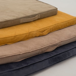 Puede incluir: Cuatro cojines de suelo cuadrados en diferentes colores y texturas. Los cojines están apilados uno encima del otro, con el cojín superior de color marrón claro, el segundo cojín de color amarillo mostaza, el tercer cojín de color marrón claro y el cojín inferior de color azul oscuro.