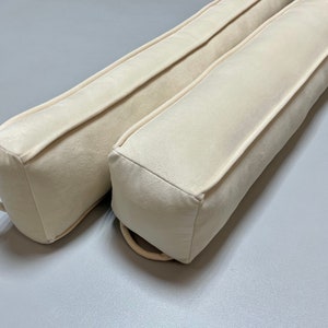 May include: Two off-white bolster pillows with a simple design and a subtle texture. The pillows are made of a soft, plush fabric and have a rectangular shape.
