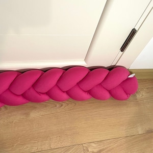 May include: A pink braided doorstop made of fabric. The doorstop is placed at the bottom of a white door.
