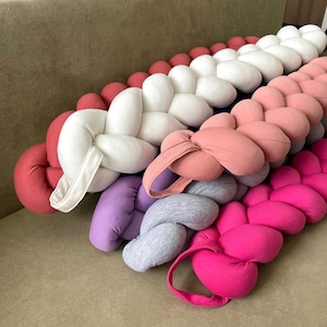 Puede incluir: Una almohada trenzada hecha de diferentes telas de colores. La almohada está hecha de seis colores diferentes: rojo, blanco, rosa, morado, gris y rosa. La almohada está trenzada de forma que crea una serie de bucles.