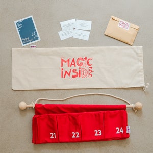 Puede incluir: Un calendario de adviento de tela roja con los n&uacute;meros blancos del 21 al 24, colgado de una varilla de madera con una cuerda. Incluye una bolsa de tela beige con el texto rojo MAGIC INSIDE, una tarjeta azul, cuatro tarjetas peque&ntilde;as y un sobre.
