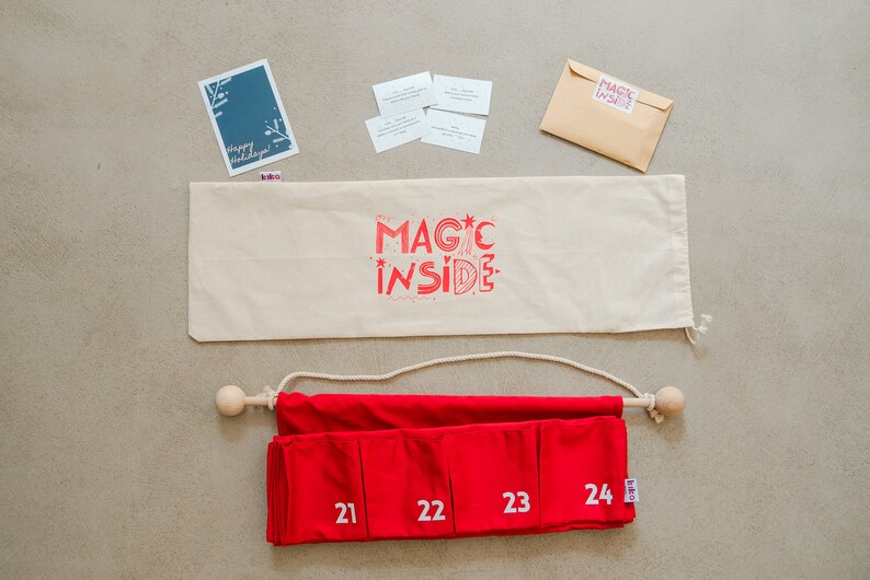 Puede incluir: Un calendario de adviento de tela roja con los n&uacute;meros blancos del 21 al 24, colgado de una varilla de madera con una cuerda. Incluye una bolsa de tela beige con el texto rojo MAGIC INSIDE, una tarjeta azul, cuatro tarjetas peque&ntilde;as y un sobre.