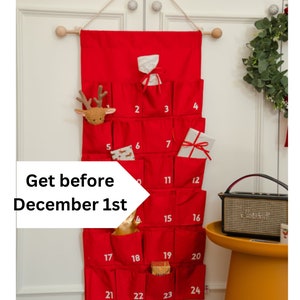 Puede incluir: Un calendario de Adviento de tela roja para colgar en la pared con veinticuatro bolsillos numerados. El calendario presenta n&uacute;meros blancos impresos del 1 al 24, con varios peque&ntilde;os regalos en algunos bolsillos, suspendido de una vara de madera.