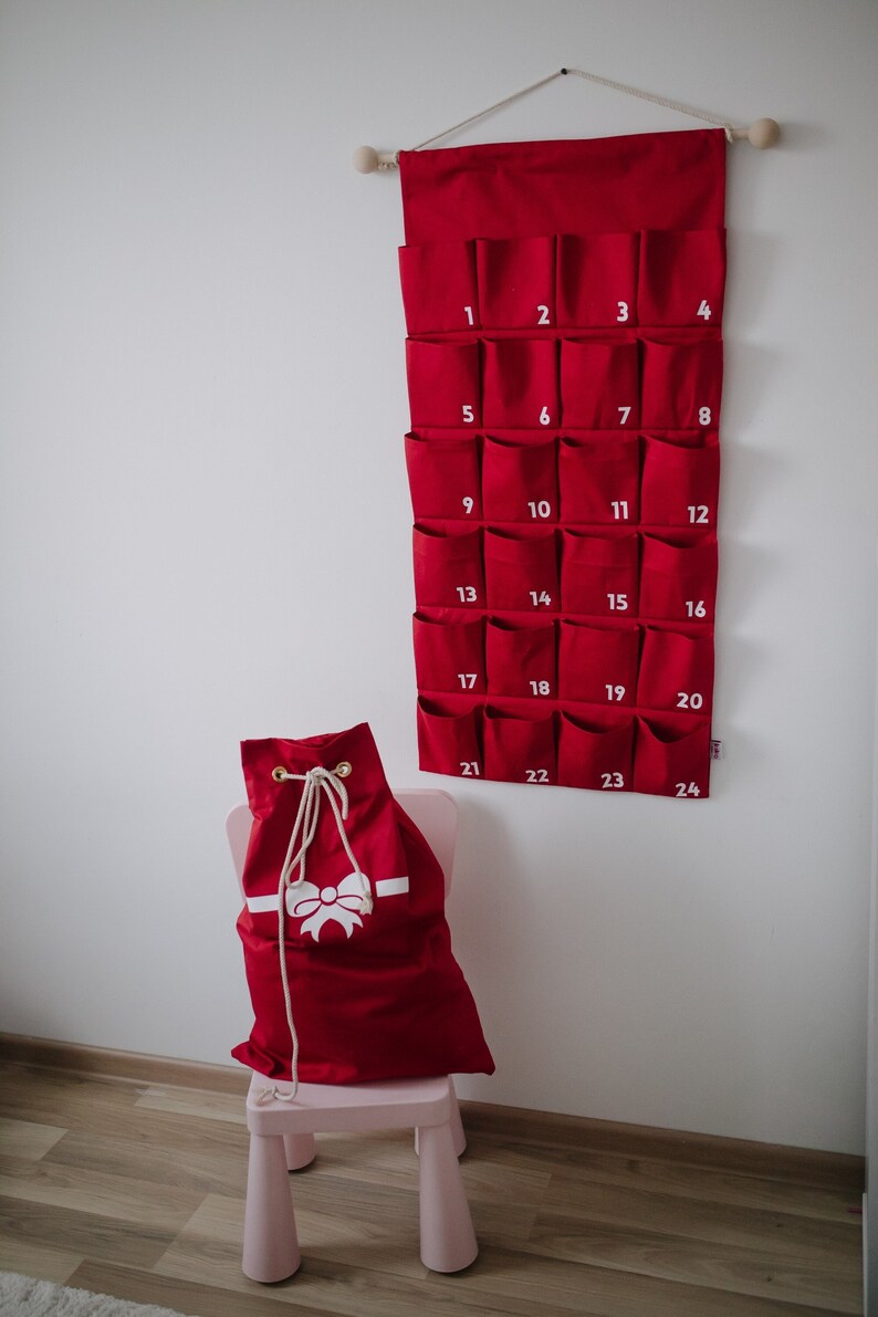 Puede incluir: Un calendario de adviento de pared hecho de tela roja con veinticuatro bolsillos numerados y un saco rojo a juego con cord&oacute;n y un gr&aacute;fico de lazo blanco. El calendario cuelga de una varilla de madera con una cuerda y el saco est&aacute; sobre una silla rosa.