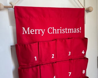 Puede incluir: Un calendario de adviento de tela roja colgado de una varilla de madera con una cuerda blanca. El calendario presenta el texto blanco Merry Christmas! e incluye bolsillos numerados del 1 al 12 visibles, dispuestos en filas sobre la tela roja.
