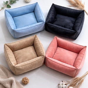 May include: Four square pet beds in pastel colors: light blue, black, tan, and pink. Each bed has a soft, plush appearance with a raised edge and a recessed center. The beds are arranged on a white surface, with decorative elements.