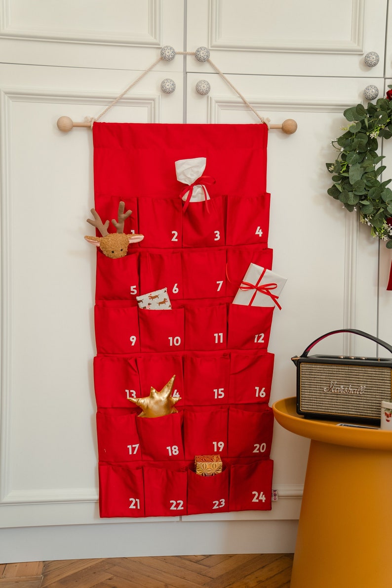 Puede incluir: Un calendario de adviento de tela roja colgado de una varilla de madera, con veinticuatro bolsillos individuales numerados en blanco. Los bolsillos contienen varios objetos peque&ntilde;os, como una cabeza de reno de peluche, una estrella dorada y regalos.