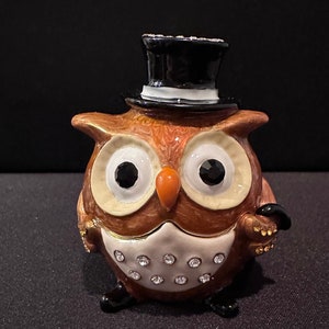 May include: A decorative owl figurine with a black top hat and rhinestone accents. The owl has large black eyes, an orange beak, and a white chest with embedded rhinestones. The owl is brown and gold in color.