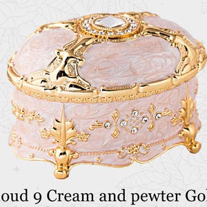 May include: Cream and gold-colored jewelry box with ornate detailing. The box features a domed lid with a central jewel and is accented with gold trim and decorative embellishments. The text "Cloud 9 Cream and pewter Gold" is at the bottom.