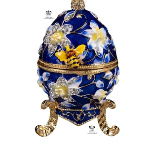 Könnte beinhalten: Dekorative blaue eiförmige Schmuckschatulle mit goldenem Sockel und Akzenten. Die Schatulle hat weiße und gelbe Blumendesigns, funkelnde Kristalle und eine gelb-schwarze Biene. Das Ei ist etwa 10 cm hoch.