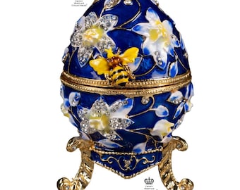 May include: A decorative blue egg-shaped trinket box with a gold base and accents. The box features white and yellow floral designs, sparkling crystals, and a yellow and black bee. The egg is approximately 4 inches tall.
