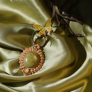 Puede incluir: Un colgante de piedra verde con un diseño tallado detallado, rodeado por un marco circular de cuentas marrones y doradas. El colgante cuelga de un cordón de cuero marrón y una mariposa amarilla está unida al cordón.
