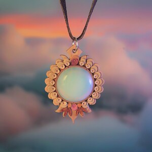 May include: A delicate necklace with a round, light blue pendant surrounded by a detailed, white, and brown floral design. The pendant is hanging against a blurred background of pink and blue clouds.