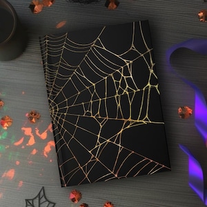 May include: A black notebook with a gold spiderweb design. The notebook is surrounded by Halloween decorations, including candles, confetti, and purple and orange ribbons. The scene is set on a gray wooden surface.