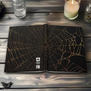 May include: Open black book with a gold spiderweb design. The book is on a dark wooden surface, with decorative butterflies and candles. The spiderweb design covers both open pages of the book.