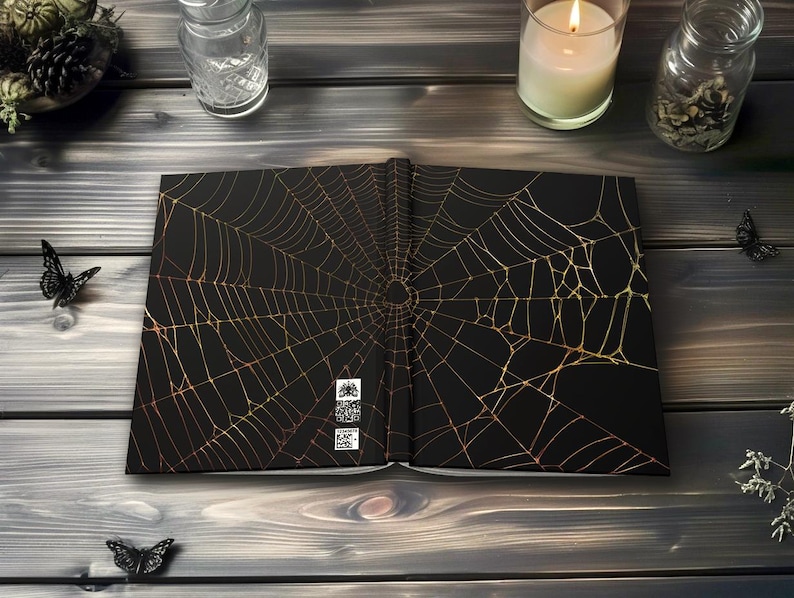 May include: Open black book with a gold spiderweb design. The book is on a dark wooden surface, with decorative butterflies and candles. The spiderweb design covers both open pages of the book.