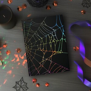 May include: A black notebook with a holographic spiderweb design. The notebook is surrounded by Halloween decorations, including candles, confetti, and a purple ribbon. The overall theme is spooky and festive.