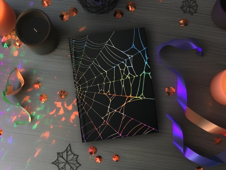 May include: A black notebook with a holographic spiderweb design. The notebook is surrounded by Halloween decorations, including candles, confetti, and a purple ribbon. The overall theme is spooky and festive.