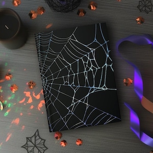 May include: A black notebook with a white spiderweb design, surrounded by Halloween decorations. The scene includes candles, confetti, and a purple and orange ribbon, creating a spooky aesthetic.