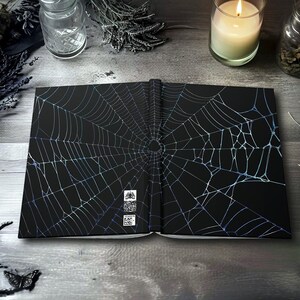 May include: Open black journal with a detailed, iridescent spiderweb design. The journal is open, revealing the inside pages. The background includes a lit candle, dried plants, and a butterfly, creating a gothic or Halloween theme.
