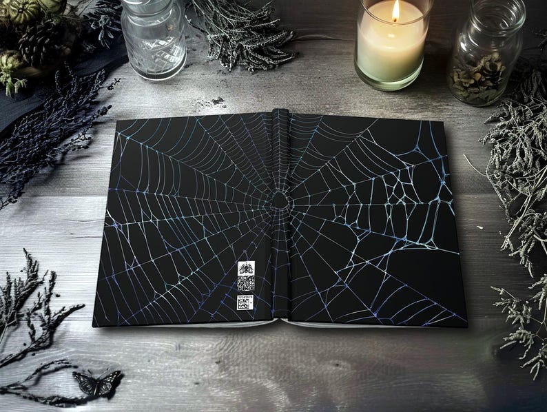 May include: Open black journal with a detailed, iridescent spiderweb design. The journal is open, revealing the inside pages. The background includes a lit candle, dried plants, and a butterfly, creating a gothic or Halloween theme.
