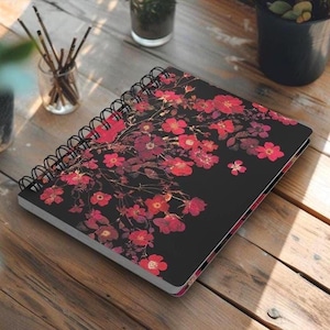 May include: A spiral-bound notebook with a black cover adorned with a floral design featuring red and pink blossoms. The notebook rests on a wooden surface, with a pen and other objects nearby.