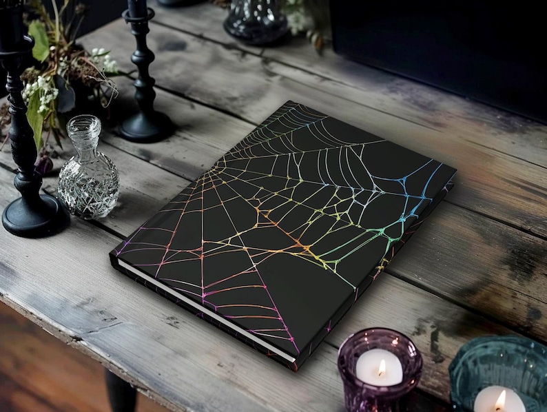 May include: A black journal with a holographic spiderweb design. The cover features a detailed web pattern with iridescent lines. The journal is set on a rustic wooden table, with candles and decorative items in the background.
