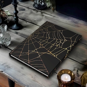 May include: A black journal with a gold spiderweb design on the cover. The journal is lying on a wooden table, with candles and other decorative items in the background. The journal has a white edge.