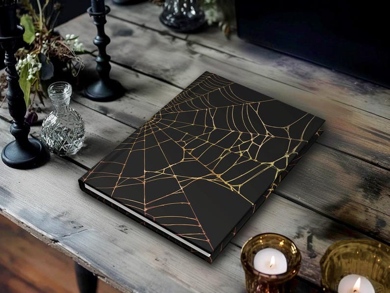 May include: A black journal with a gold spiderweb design on the cover. The journal is lying on a wooden table, with candles and other decorative items in the background. The journal has a white edge.