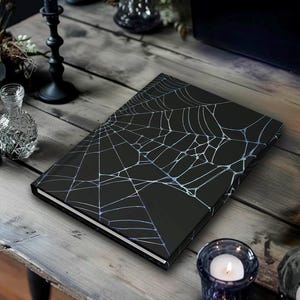 May include: A black journal with a white and light blue spiderweb design. The journal is on a wooden table, with candles and decorative items in the background. The journal is a perfect gift for Halloween.