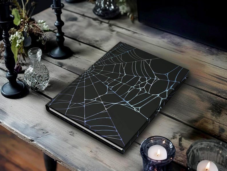 May include: A black journal with a white and light blue spiderweb design. The journal is on a wooden table, with candles and decorative items in the background. The journal is a perfect gift for Halloween.