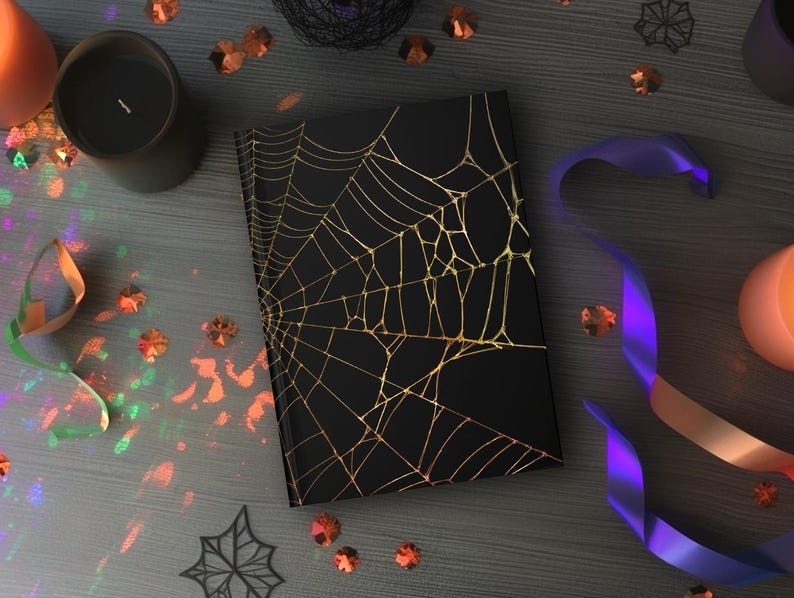 May include: A black notebook with a gold spiderweb design. The notebook is surrounded by Halloween decorations, including candles, confetti, and purple and orange ribbons. The scene is set on a gray wooden surface.