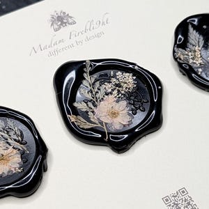 May include: Three black wax seals with dried flowers and foliage encased within. The seals have a glossy finish and are arranged on a white surface. The text "Madam Firelight different by design" is visible.